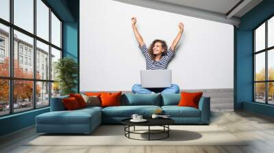 Portrait of happy young mixed races curly woman sitting on floor with laptop and celebrate win Wall mural